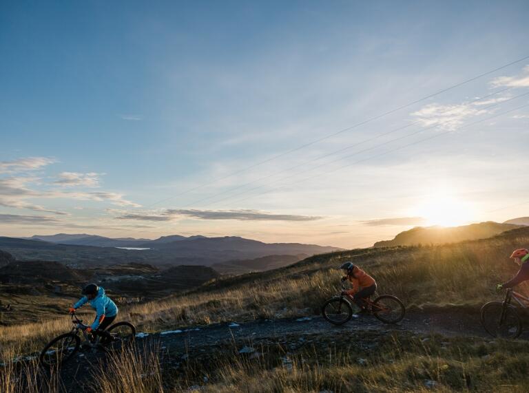 Three people on mountain bikes riding along a single track mountain trail with mountain scenery and a low sun in the sky in the background.