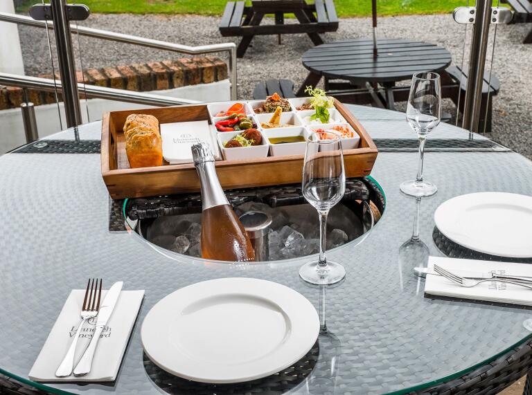 A selection of food in a wooden tray with a loaf of bread set of a table laid out with crockery and wine.