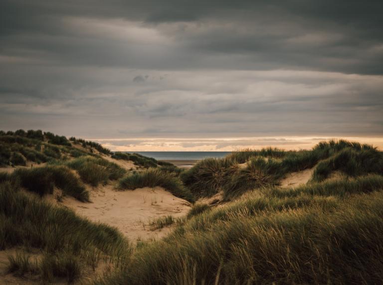 Distant sea views between grass-topped sand dunes under dark skies.