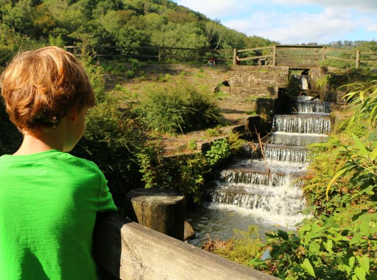 Ein Junge in einem grünen T-Shirt schaut mit dem Rücken zur Kamera auf einen Wasserfall.