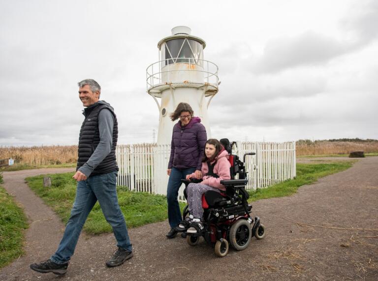 Girl in wheelchair with parents on a path in front of a lighthouse