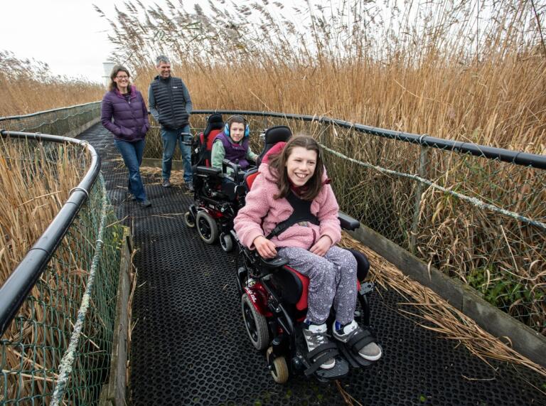 Two children in wheelchairs with their parents on a path with reeds on either side
