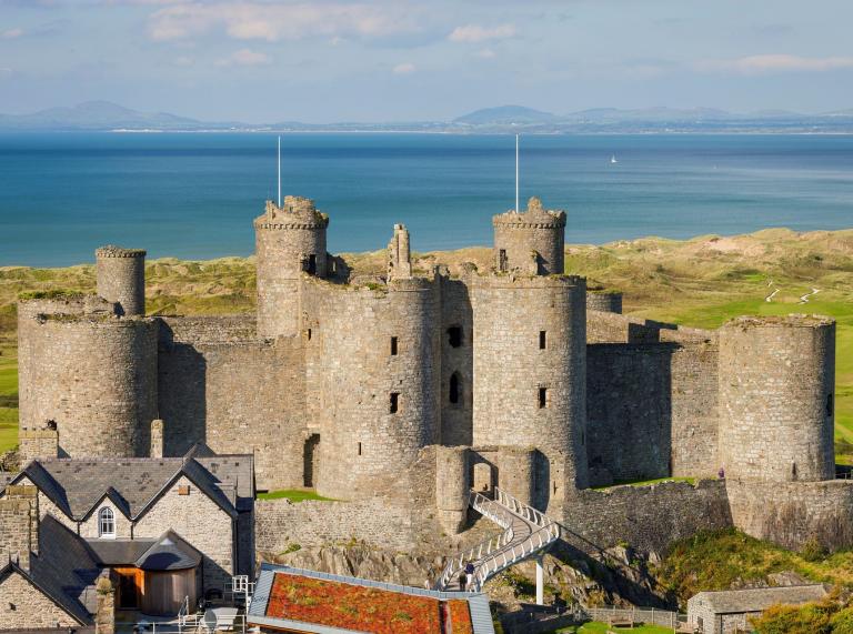 A view of Harlech Castle and its white walkway leading to the entrance with the sea and golf course beyond, 