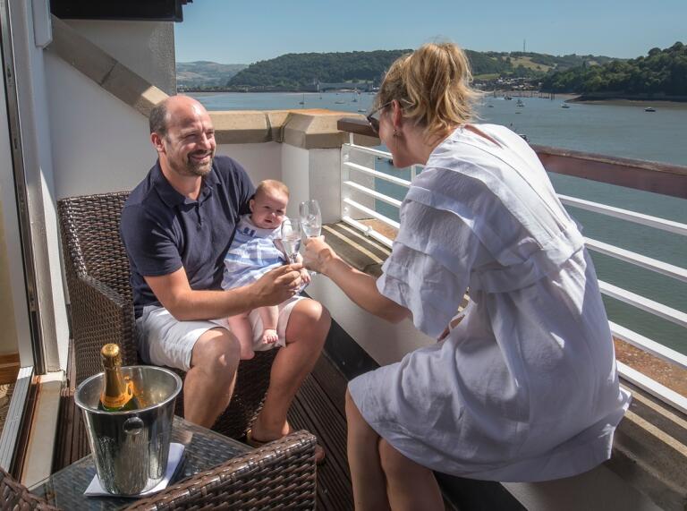 Ein Paar mit kleinem Baby genießt ein Glas Wein auf einem Hotelbalkon mit Blick auf den Fluss.