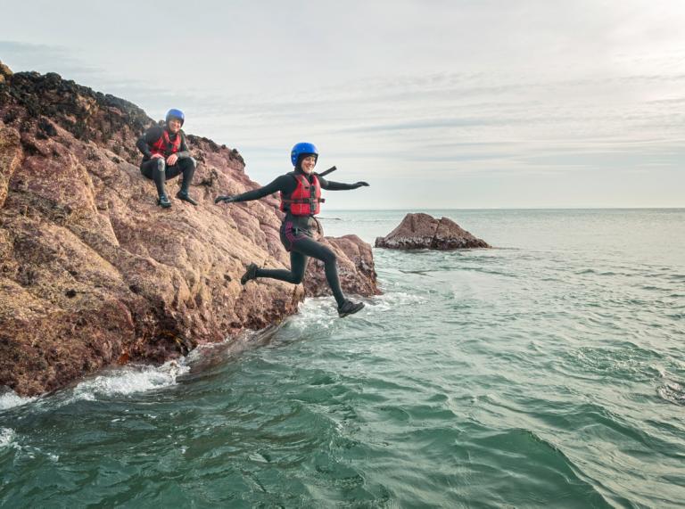 A lady jumping off a rock into the sea wearing safety gear with a friend looking on.