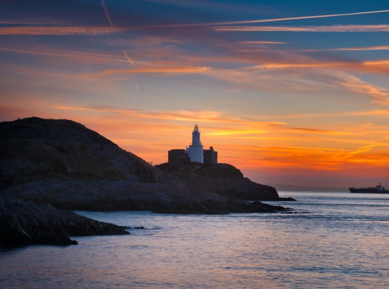 A lighthouse on the edge of a rocky headland against a backdrop of an orange sunrise.