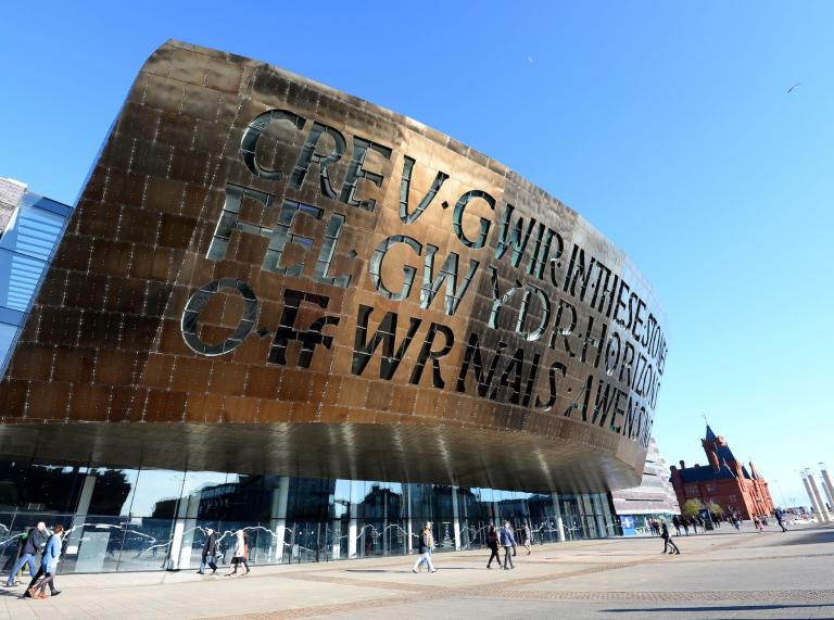 People walkin outside Wales Millennium Centre with the sunlight reflected on the brass carving.