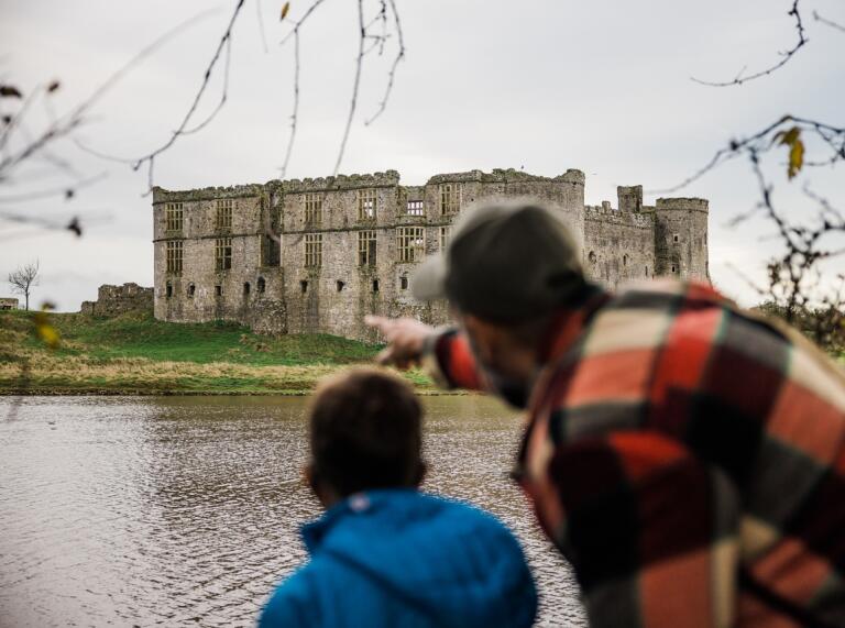 Mann und Junge blicken zum Carew Castle mit der Flussmündung des Cleddau im Vordergrund. Der Mann zeigt auf die Burg.
