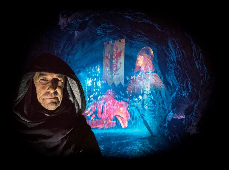 A hooded monk with a magical waterfall behind him in an underground cave.