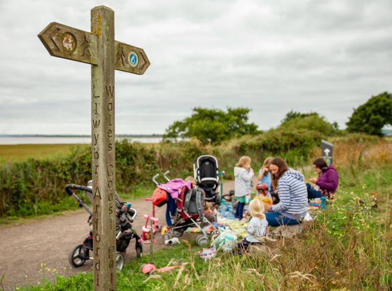 A group of parents and young children with pushchairs having a break by a Wales Coast Path sign.