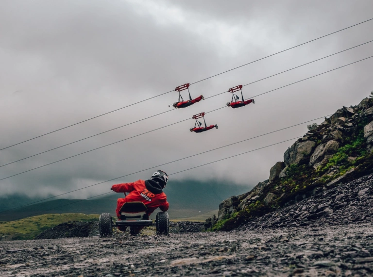 A go carter driving down a quarry with people passing by on zip lines.