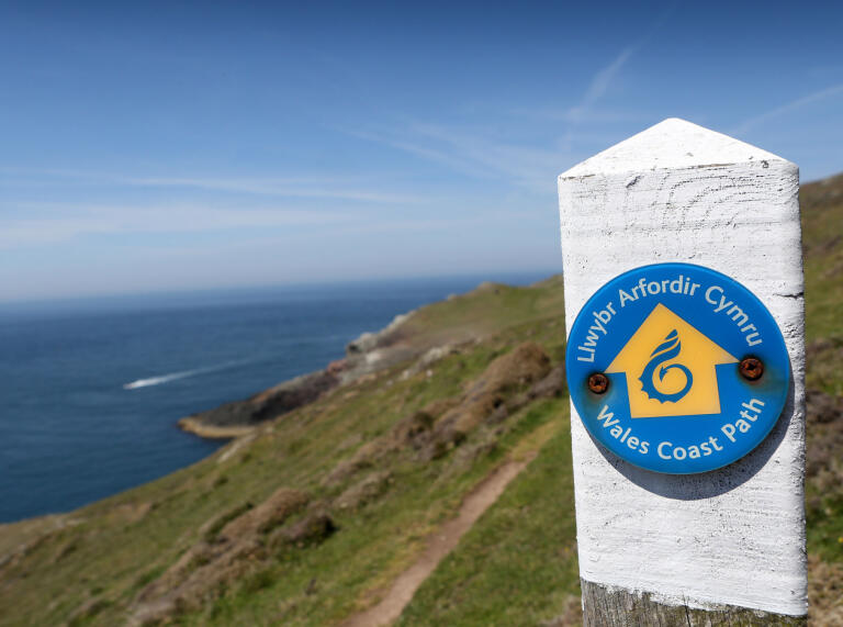 Ein weißer Pfosten mit einer blauen Plakette zeigt Wanderern den Weg auf dem Wales Coast Path.