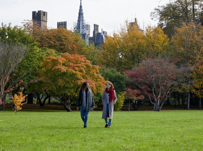 Zwei Frauen bei einem Spaziergang im Bute Park mit herbstlichen Bäumen und Cardiff Castle im Hintergrund.