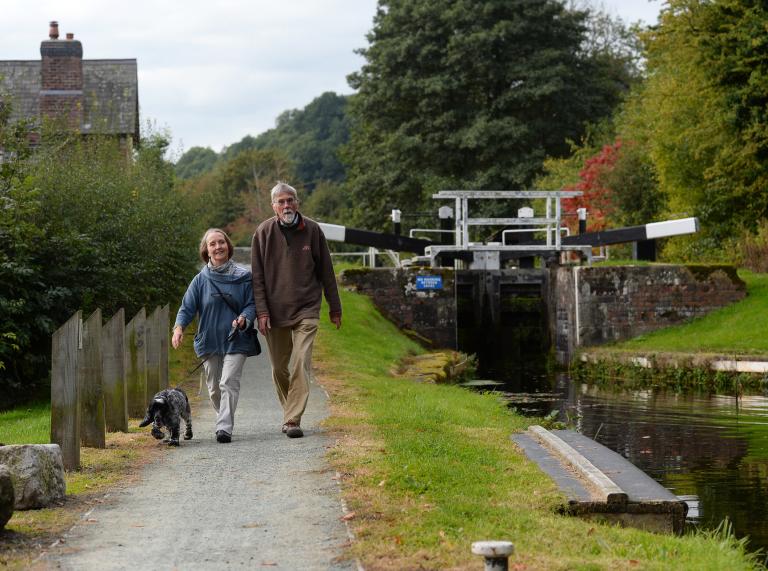 A woman, a man and a dog walking alongside a canal with a lock in the background.