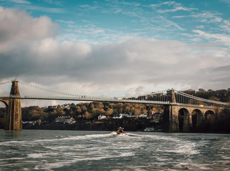 Image of the suspension bridge with houses and trees in the distance, and a rib boat on the Menai Strait