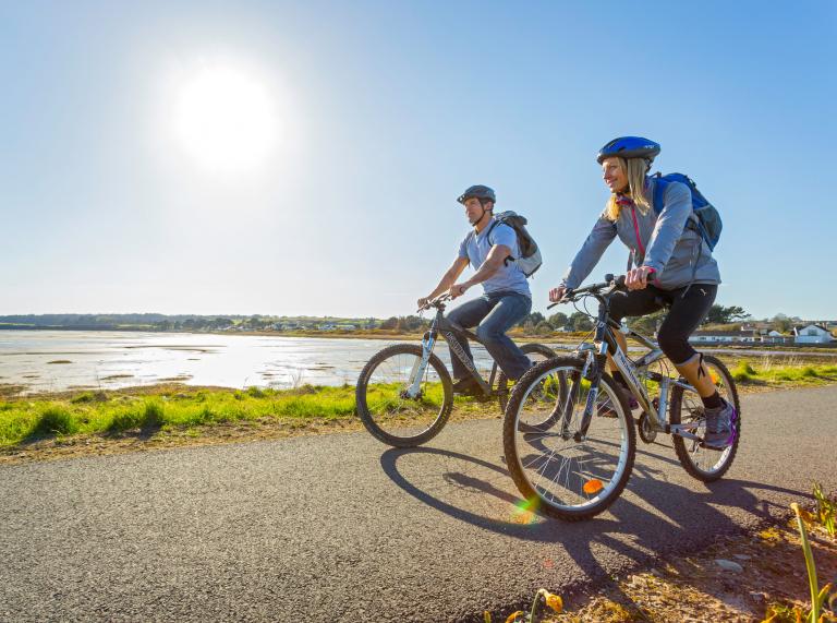 Two people cycling on a coastal cycle path on a sunny day.