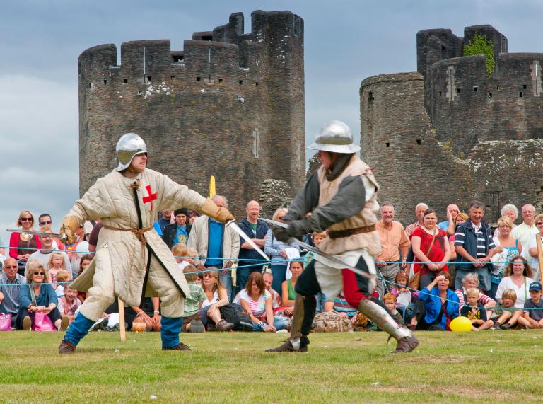 Two men dressed in medieval costumes taking part in a battle re-enactment, with crowd watching by the castle.