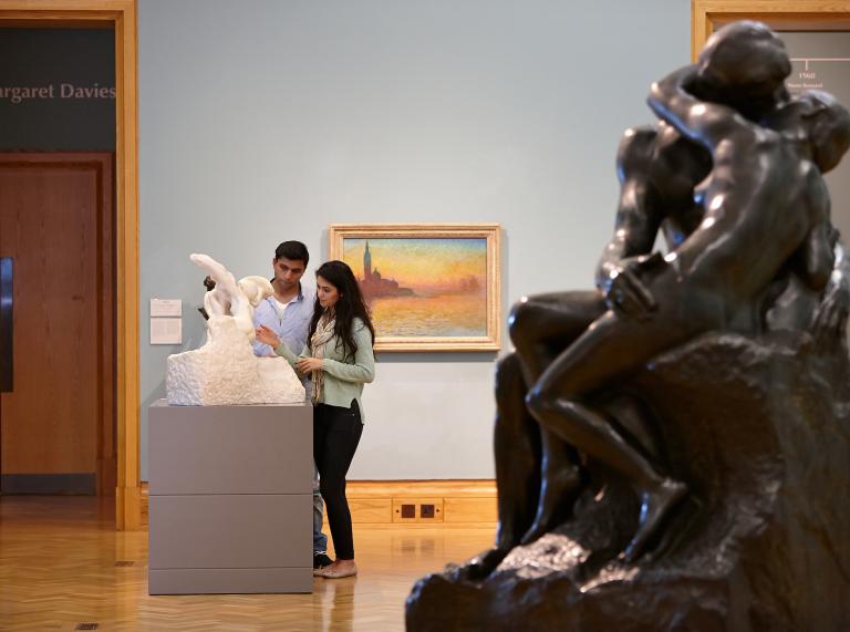 A couple looking at a small statue, with a painting behind them, and a large sculpture to the side in the foreground