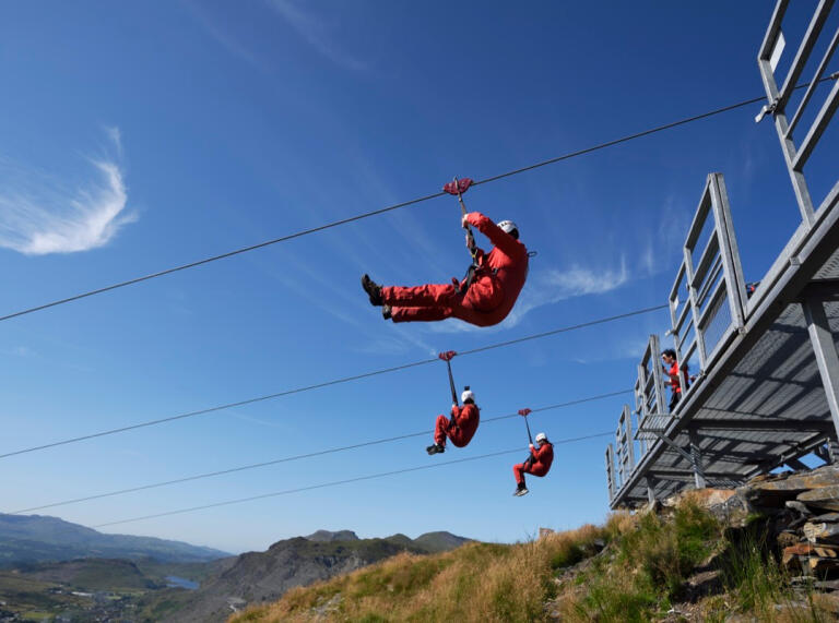 Three people sitting in a harness on parallel zip lines.