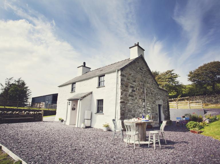 exterior view of white stone cottage with gravel out front and table and chairs and fences and field in the background