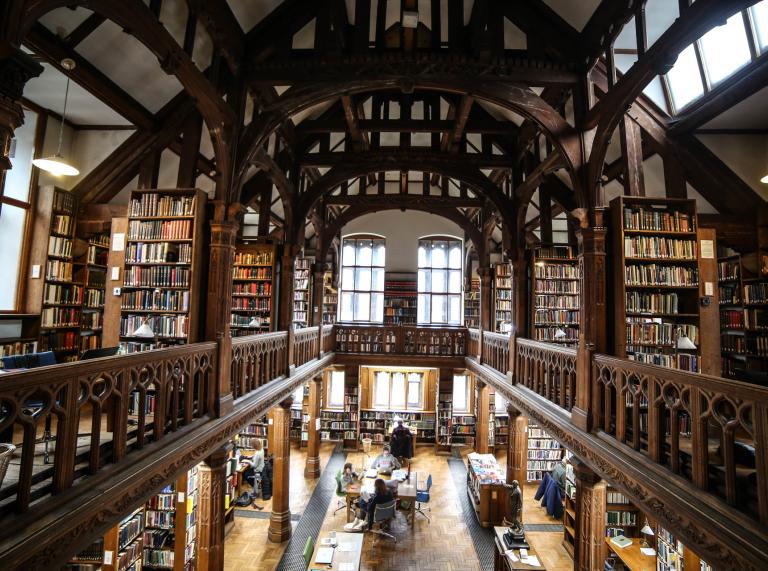 Innenansicht einer Bibliothek mit hoher Decke und Holzregalen und Menschen, die an Schreibtischen im Erdgeschoss sitzen.