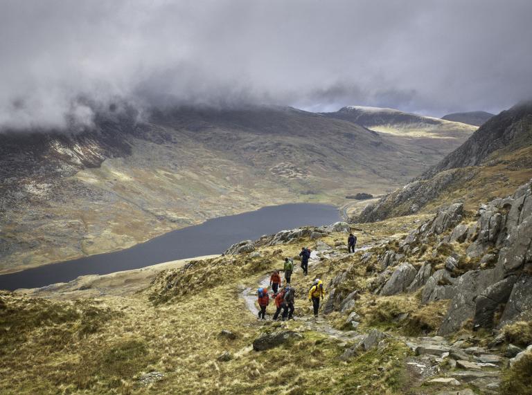 Wanderer an einem Berg auf dem Weg zum Gipfel mit einem Bergsee im Hintergrund und tiefhängenden Wolken.