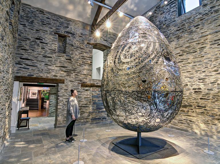Giant egg sculpture with woman stood looking at it inside a stone wall room.