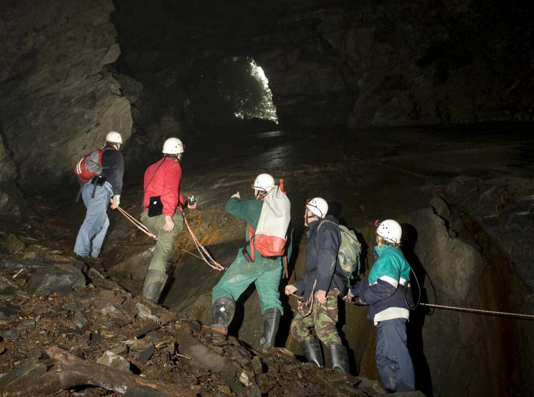 people with hard hats with lights on exploring mine with shaft of light coming through and rope being held