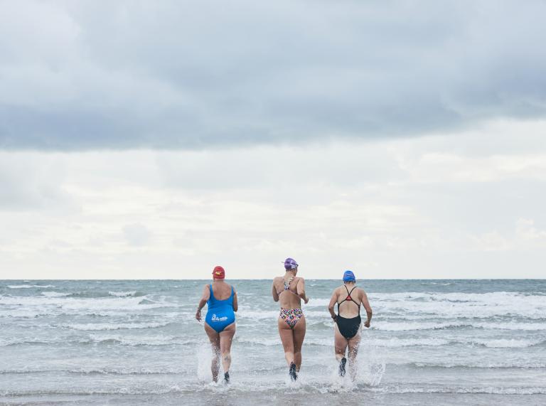 Drei Damen in Badeanzügen laufen ins Meer.