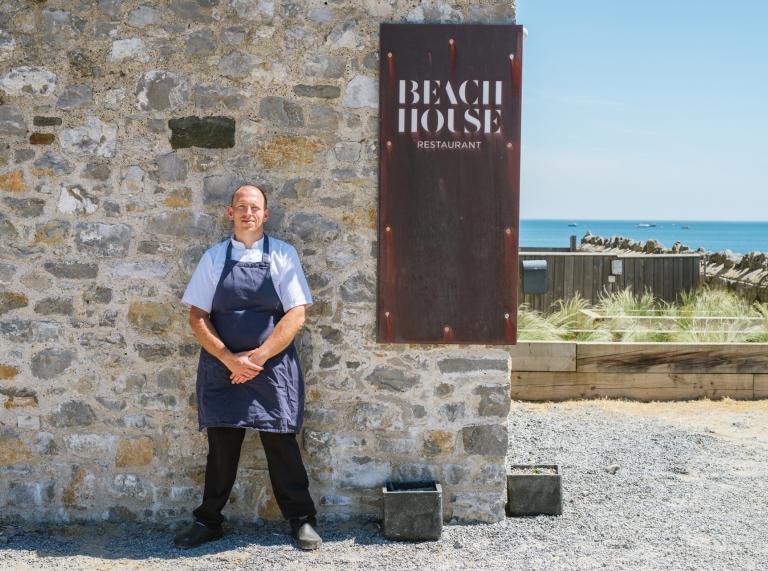 Der Chefkoch steht vor seinem Restaurant mit dem Strand im Hintergrund.