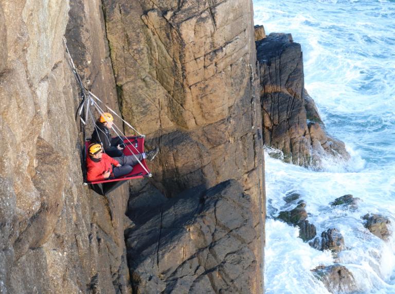 A cliff face above the sea with two people sitting on a portaledge suspended from the cliff edge.