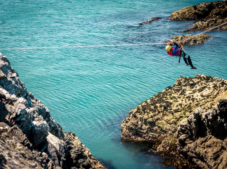 man in safety equipment under high wire moving from rocks over the blue waters.