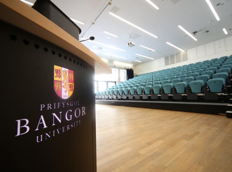 Auditarium with blue chairs and Bangor University logo and name on a black podium. 