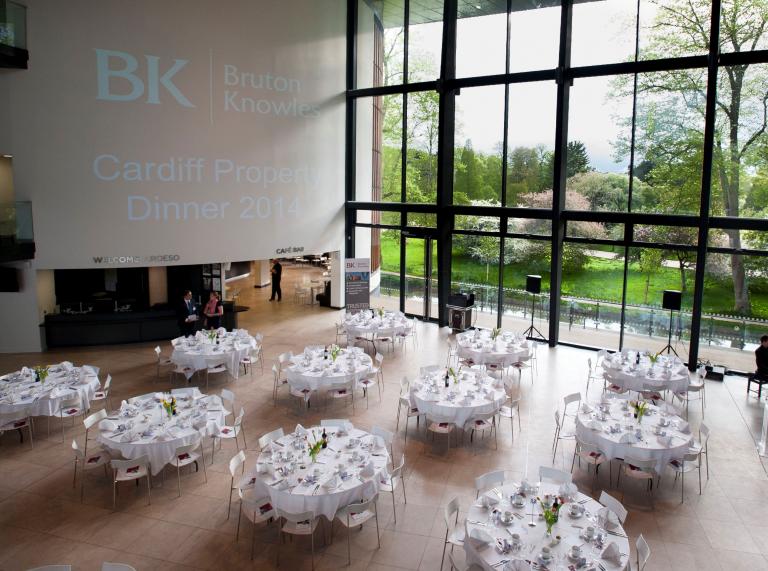 Hall set up with round dining tables, looking out onto green parkland with floor to ceiling windows. 