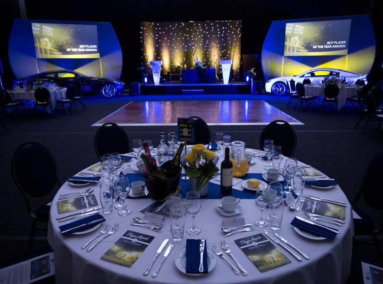 Table set for awards ceremony with white table cloth, yellow flowers, crockery on table and lit stage.