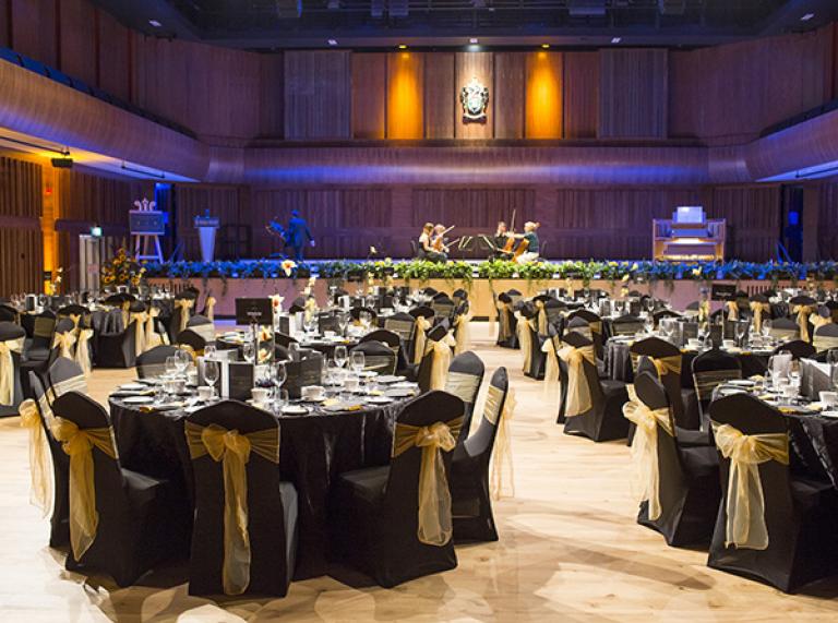Great Hall set up for a gala dinner with gold bows around black dressed chairs tables with crockery on and a stage with musicians playing.