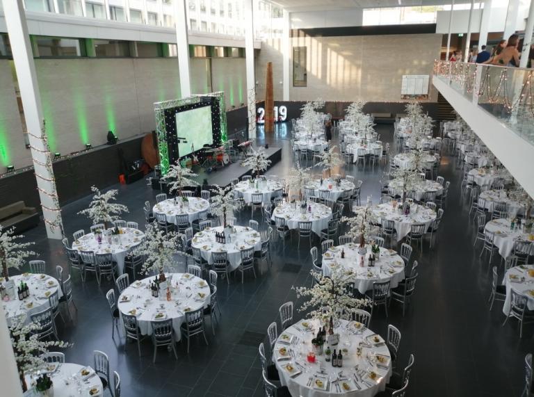 Atrium with round tables and chairs and tall white flower dress coverings , stage with fairy lights across the back.