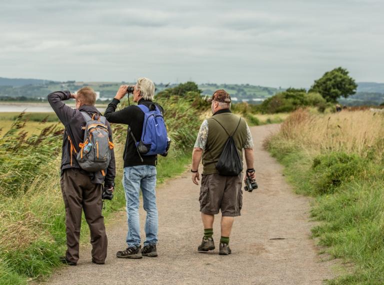 three males stood on a footpath. Two are bird watching, using binoculars to look in to the sky.