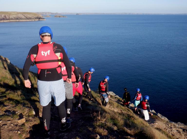 Coasteers wearing protective gear walking down a cliff toward the ocean.