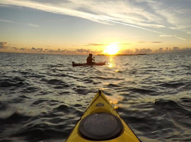 The front of a  yellow Kayak facing a person in another kayak with the sun setting in the background.