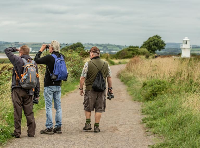 three men walking along path in wetlands, one with binoculars.