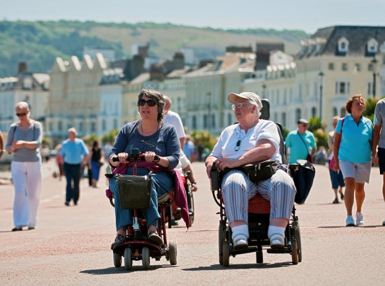 Zwei Frauen in einem Rollstuhl und einem Scooter auf der Strandpromenade.