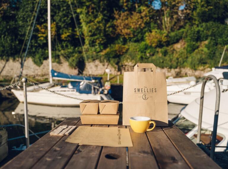 A brown paper bag with a Swellies logo, three food boxes and a yellow coffee mug on a wooden table with a boat on the water in the background