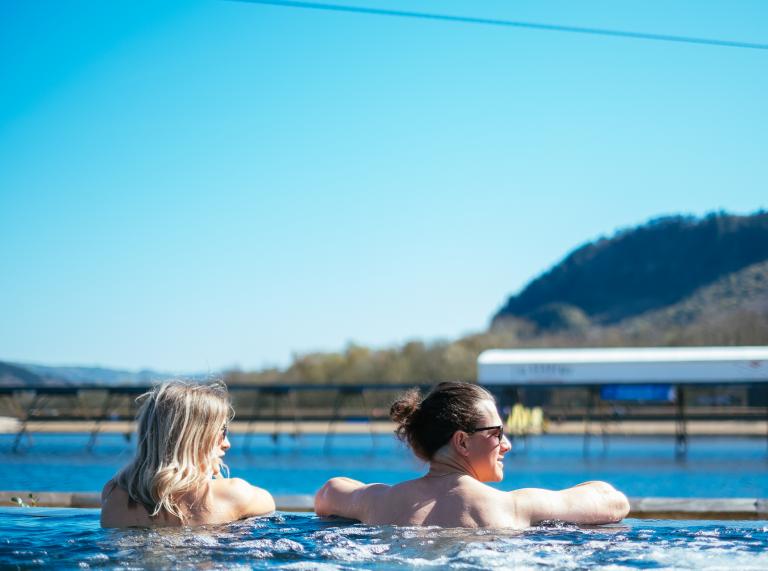Two people in an outdoor infinity pool overlooking a surf lagoon with blue skies.