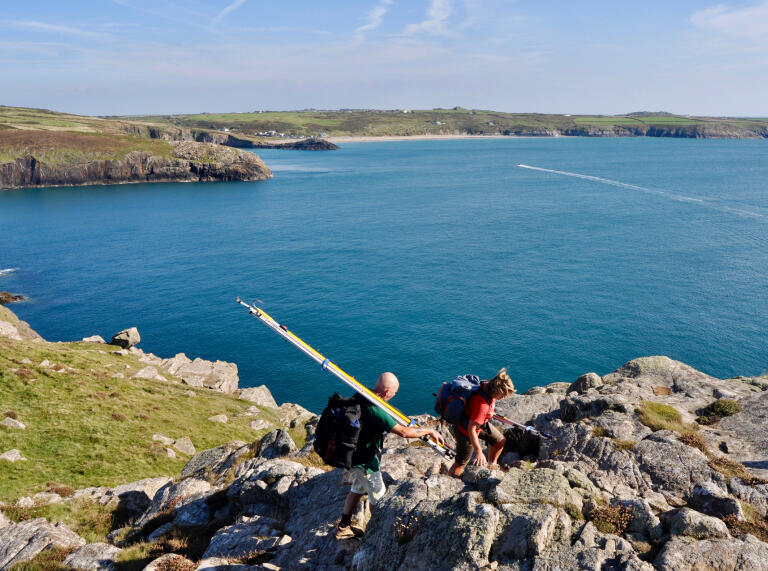 Zwei Personen mit Angeln die über Felsen klettern, mit dem Meer im Hintergrund.