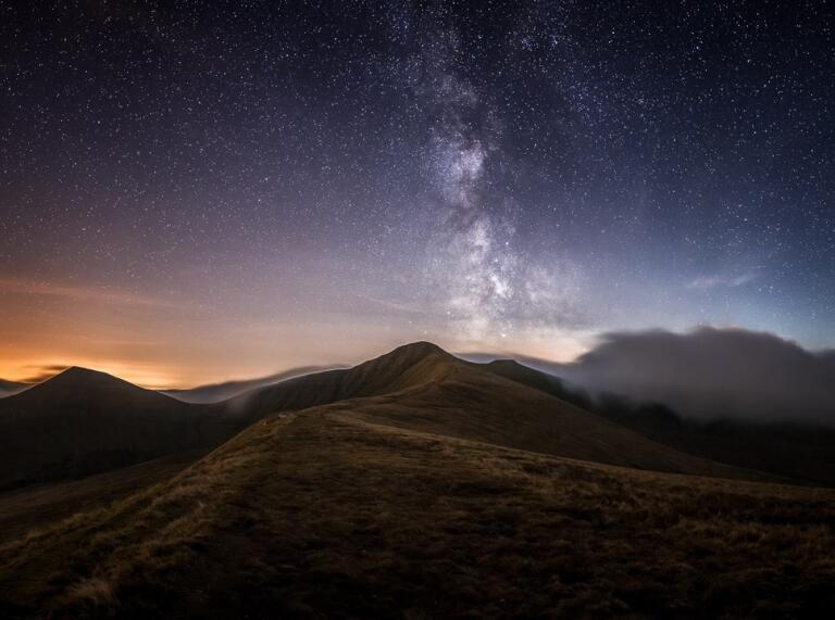 Dark sky full of stars above the mountain landscape of the Bannau Brycheiniog (Brecon Beacons) with the orange glow of the setting sun on the horizon.