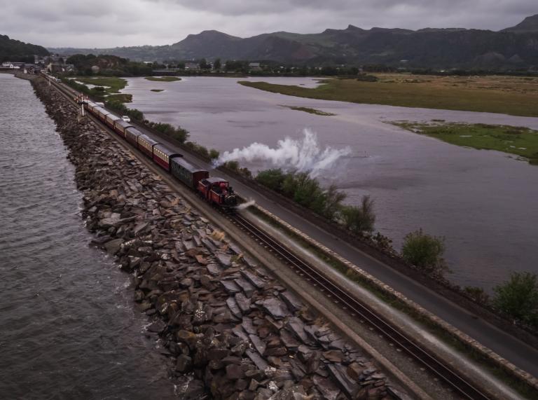 Ein Zug, der über einen Damm bei Porthmadog, umgeben von Wasser, fährt, mit Bergen im Hintergrund.