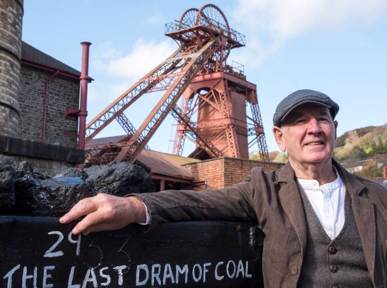 A former miner smiling at the camera with a tower colliery behind him.