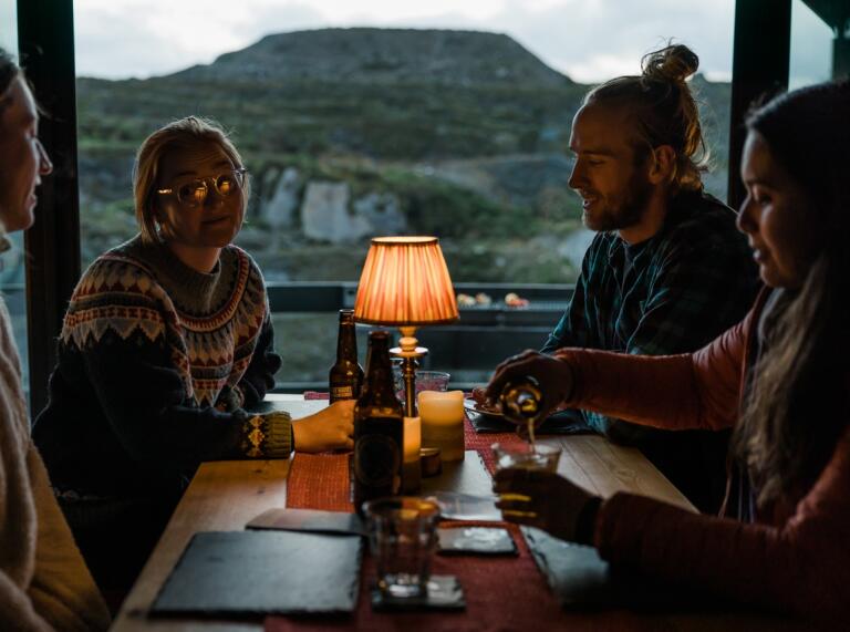 Vier Freunde am Tisch in einer der Glamping-Hütten. Auf dem Tisch steht eine Lampe, Bierflaschen und Gläser. Im Hintergrund kann man die Berge sehen.