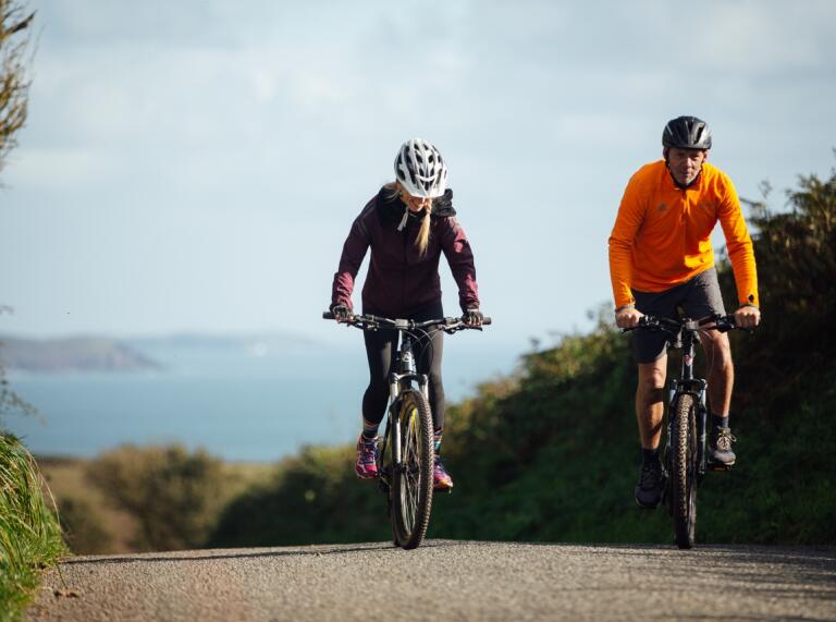 Two rider on mountain bikes riding on a coutry lane with hedgerows to each side and the sea in the far background